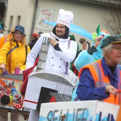 Bunt kostümierte Jecken tragen kleine Zug-Waggons wie Bauchläden mit sich. Der Narr mit dem „Bordrestaurant“ trägt eine Kochmütze.