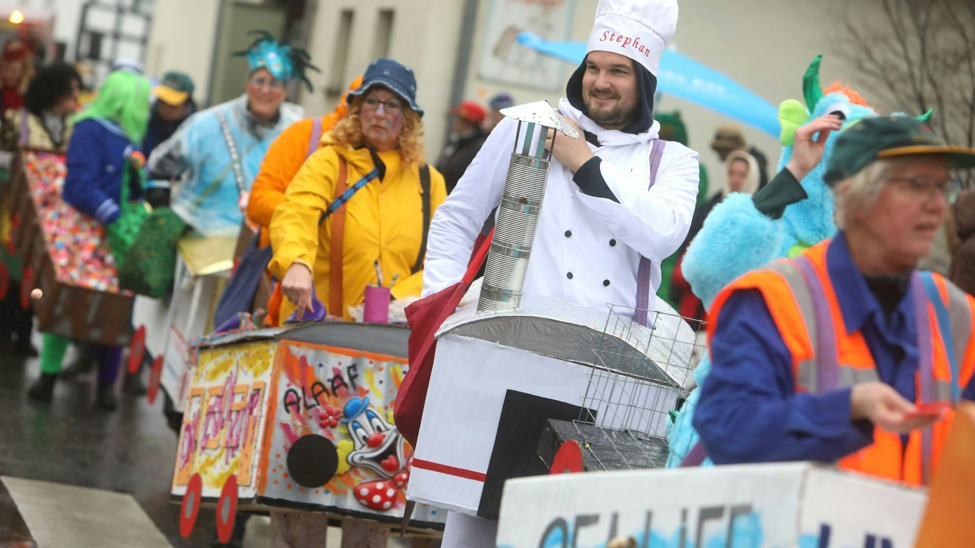 Bunt kostümierte Jecken tragen kleine Zug-Waggons wie Bauchläden mit sich. Der Narr mit dem „Bordrestaurant“ trägt eine Kochmütze.