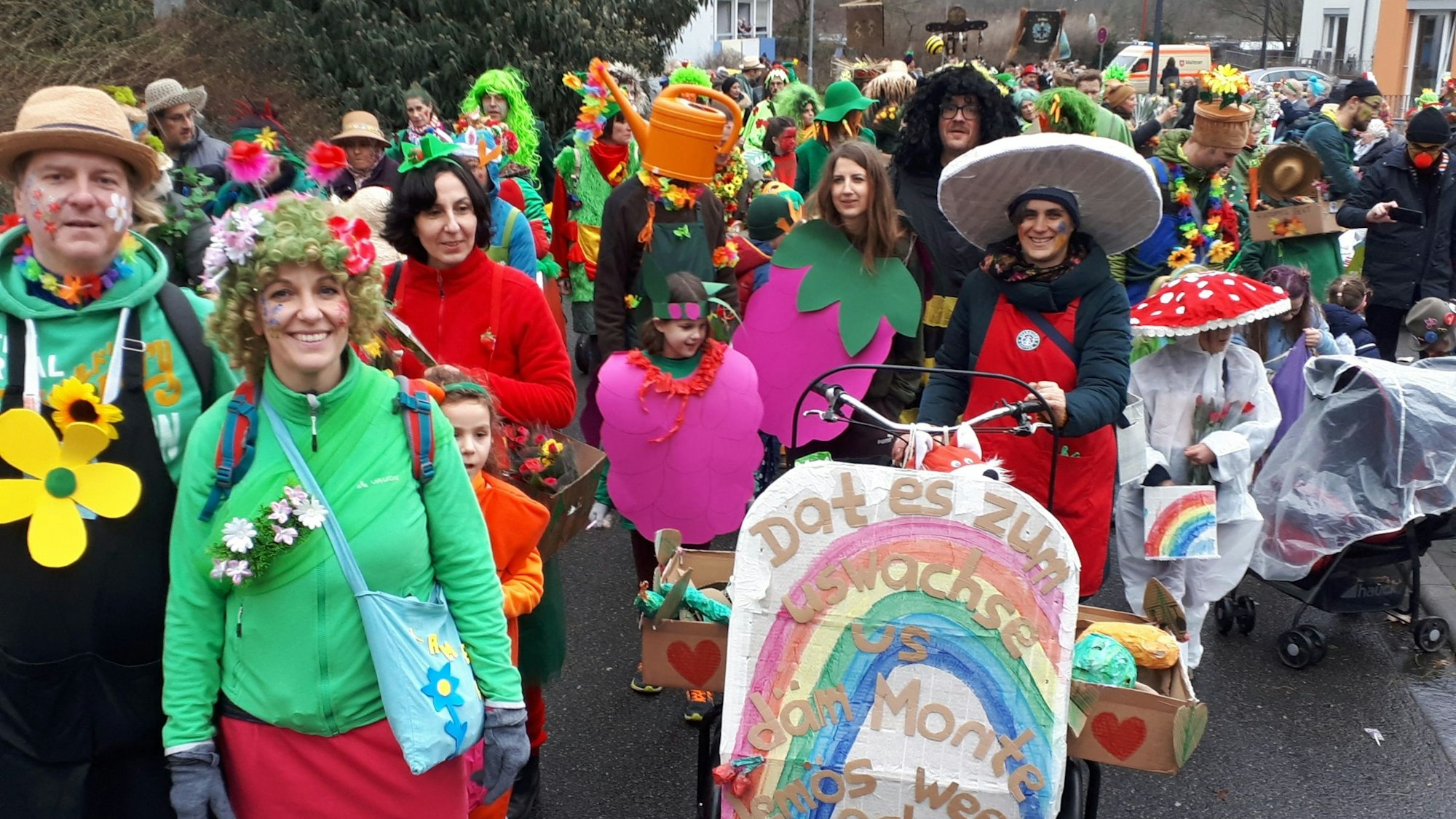 Teilnehmerinnen und Teilnehmer des Veedelszugs in Riehl stehen verkleidet auf einer Straße. Vor ihnen steht ein Schild mit der Aufschrift: „Dat es zum Uswachse – us däm Monte-Jemös weed och noch wat“.