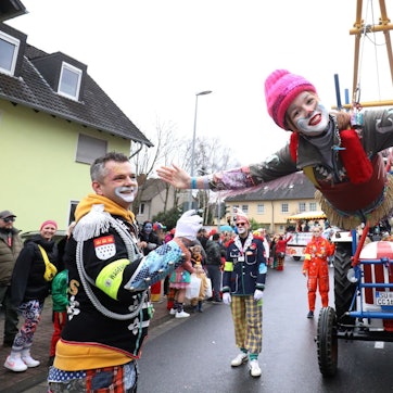 Eine bunte kostümierte Clownin schwebt als fliegender Clown an einer speziellen Konstruktion eines Traktors. Am Straßenrand stehen viele ebenfalls kostümierte Zugbesucher.