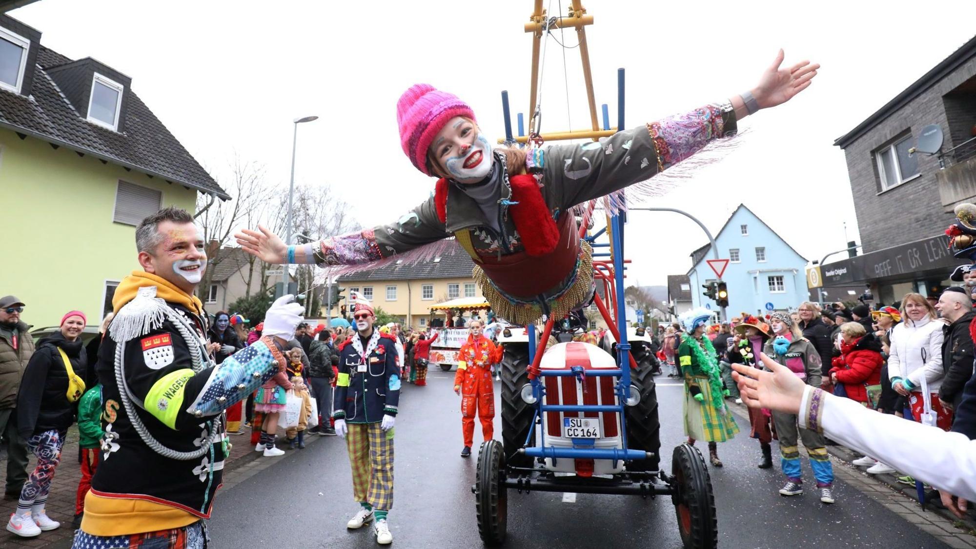 Eine bunte kostümierte Clownin schwebt als fliegender Clown an einer speziellen Konstruktion eines Traktors. Am Straßenrand stehen viele ebenfalls kostümierte Zugbesucher.