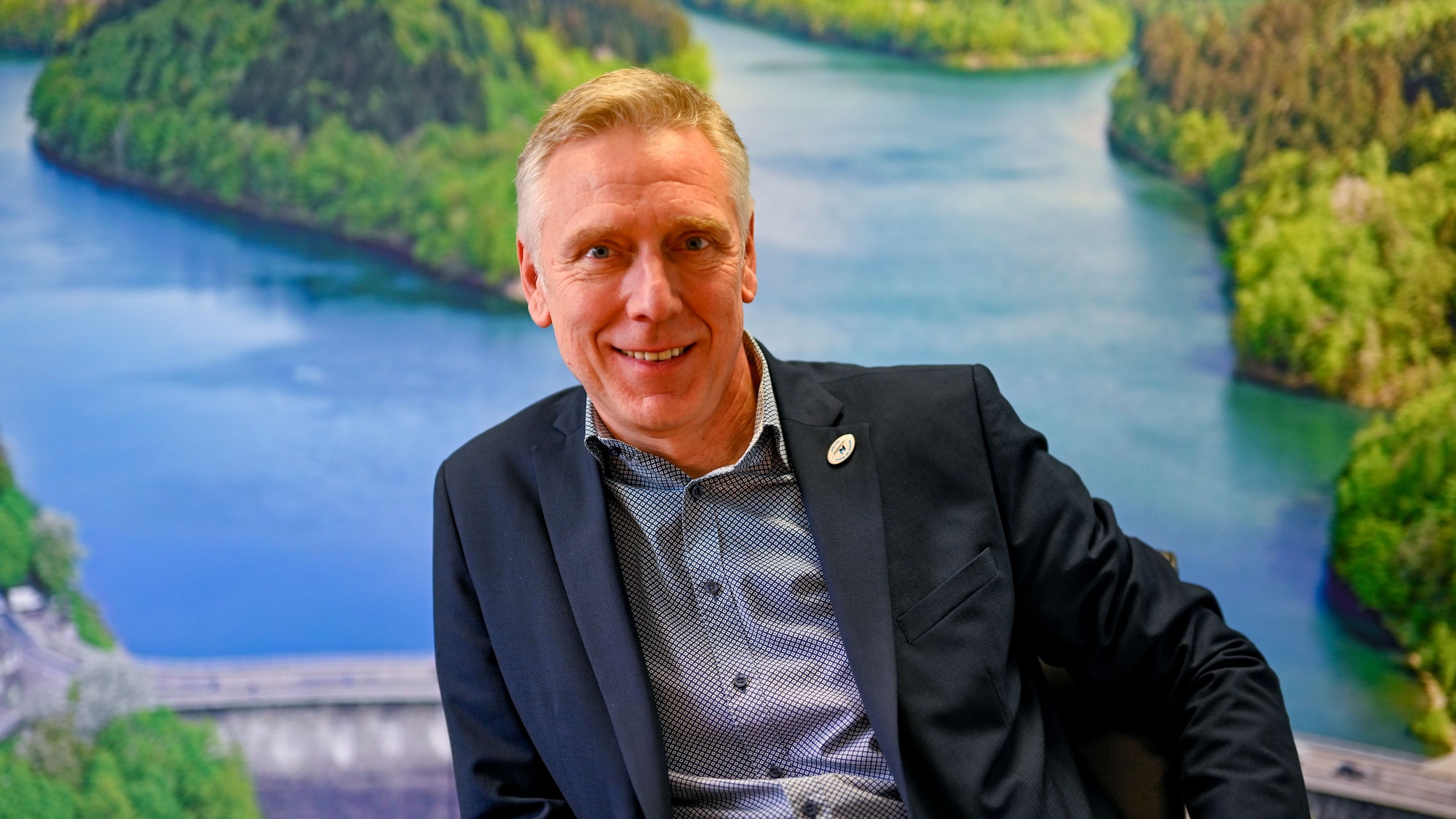 Dr. Uwe Moshage sitzt vor einer großen Luftaufnahme der Aggertalsperre.