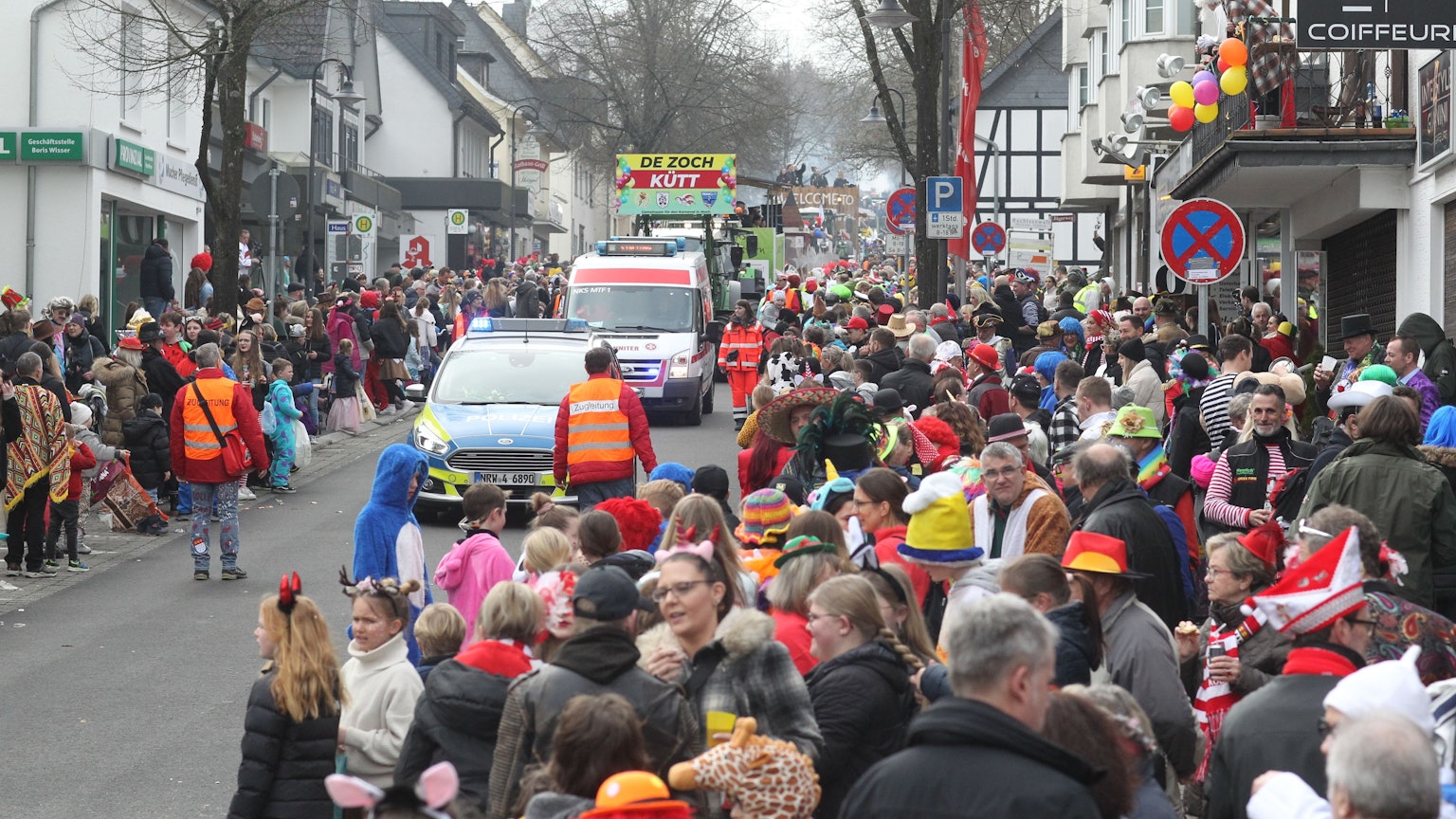 Menschen warten am Straßenrand auf einen Karnevalszug. Im Hintergrund ist ein Transparent zu sehen "De Zoch kütt" steht darauf, vorneweg fährt ein Polizeiauto.