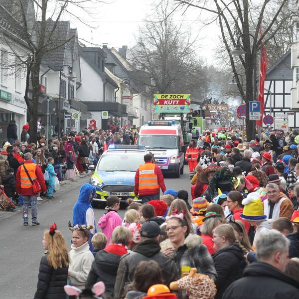 Menschen warten am Straßenrand auf einen Karnevalszug. Im Hintergrund ist ein Transparent zu sehen "De Zoch kütt" steht darauf, vorneweg fährt ein Polizeiauto.