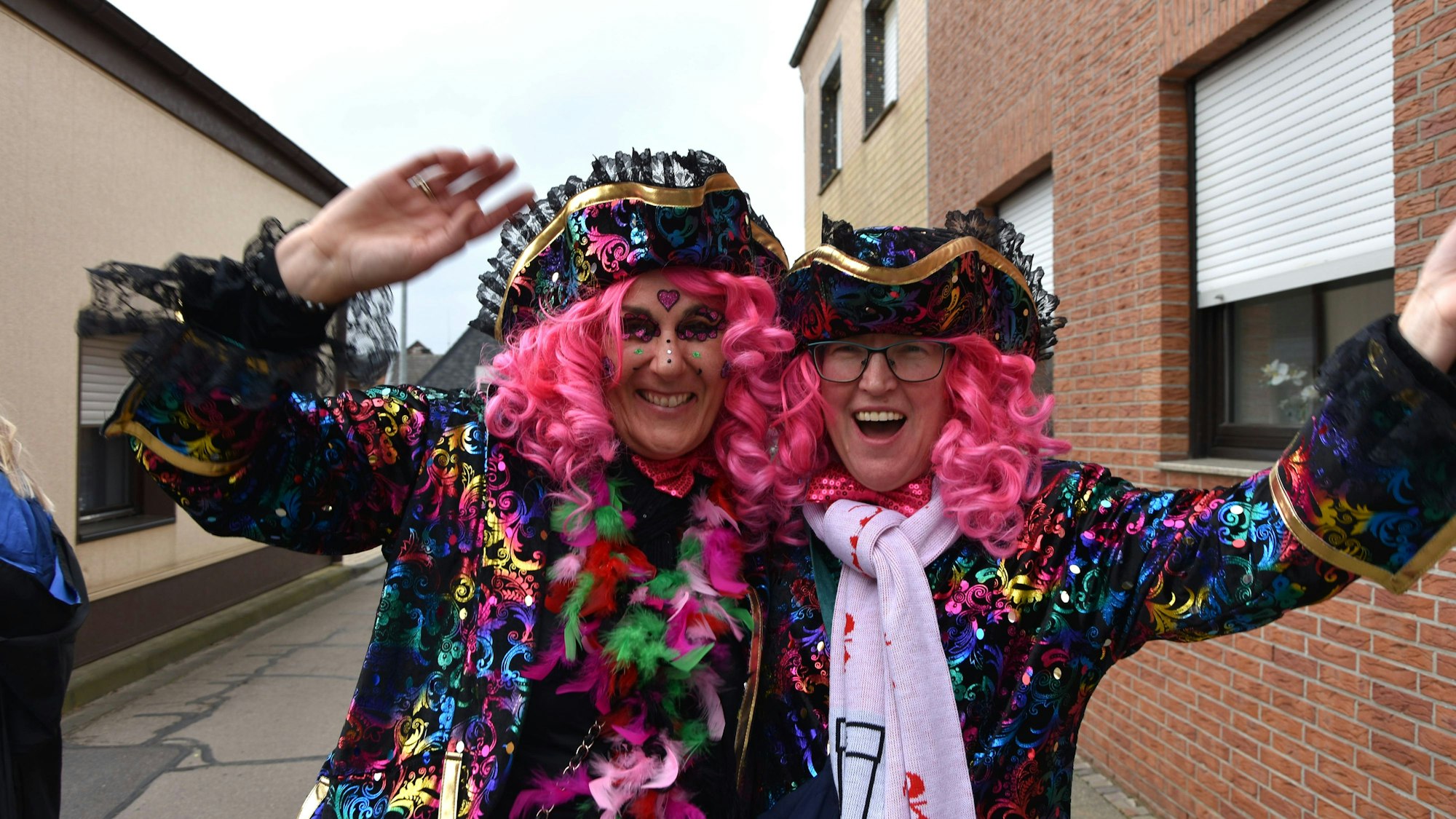 Zwei bunt kostümierte Jecken mit pinken Perücken winken in die Kamera.