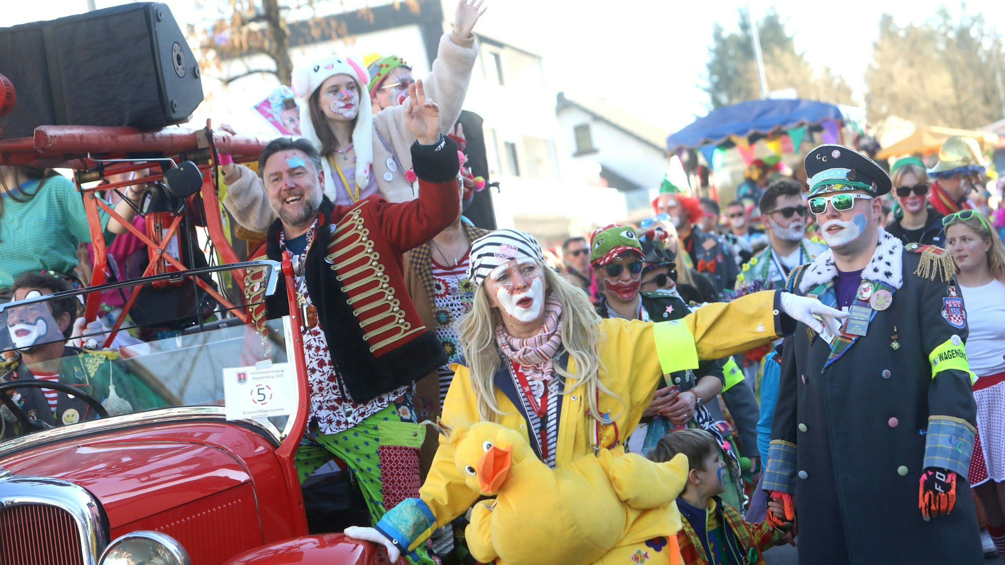 Fröhliche und bunt gekleidete Clowns ziehen durch die Straße, im Vordergrund trägt eine Närrin eine große gelbe, wohl aus Stoff gemachte Ente vor ihrem Bauch.