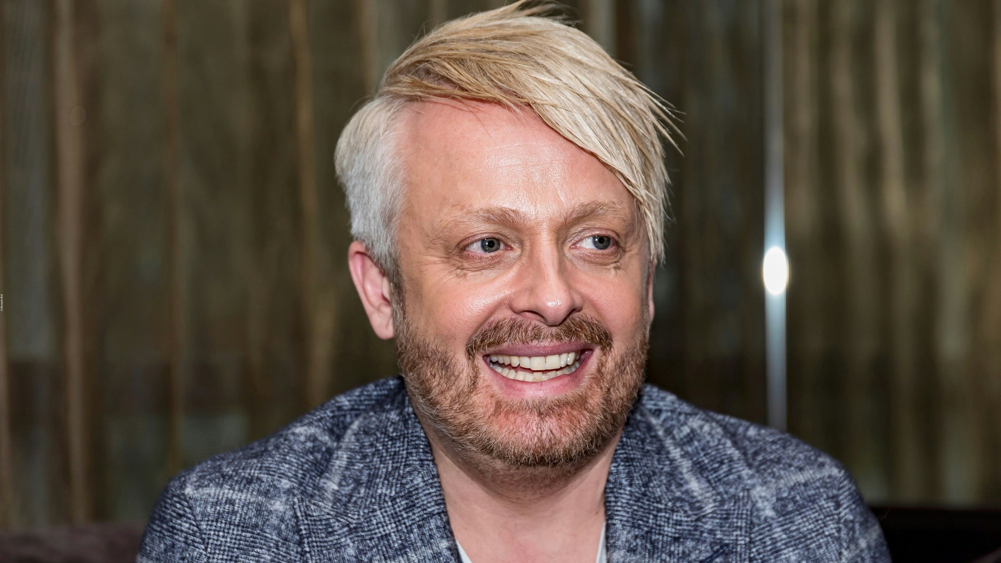 Ross Antony hat ein blau-weiß kariertes Sacko an und lacht an der Kamera vorbei.