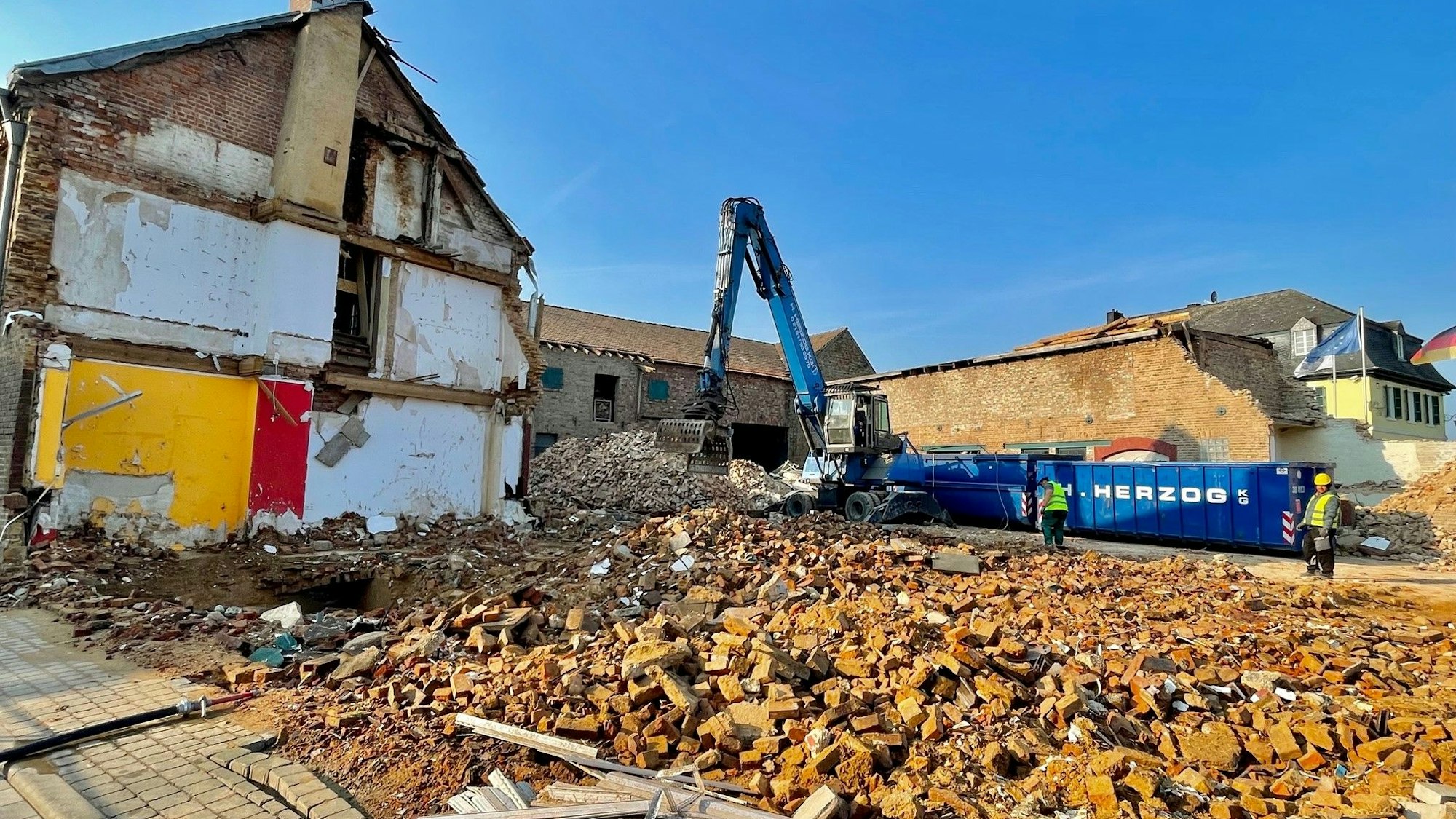 Von einem Haus sind nur noch viele Steine über, ein Bagger steht im Hintergrund, ein halb abgerissenes Haus steht an der Seite.