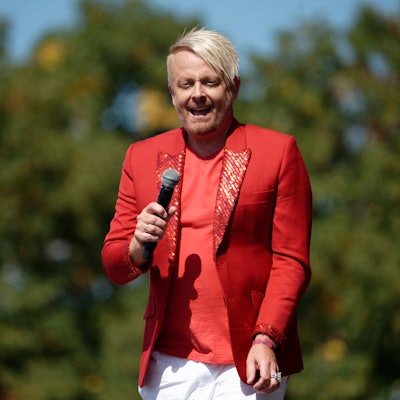 Ross Antony trägt ein rotes Sakko mit Glitzer, er hat ein Mikro in der Hand. Im Hintergrund sind Laubbäume zu erkennen.