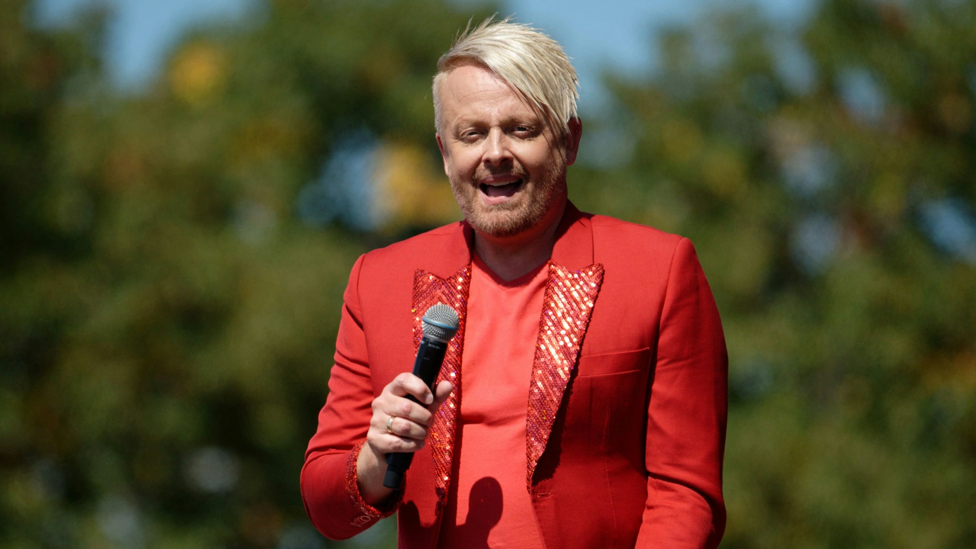 Ross Antony trägt ein rotes Sakko mit Glitzer, er hat ein Mikro in der Hand. Im Hintergrund sind Laubbäume zu erkennen.
