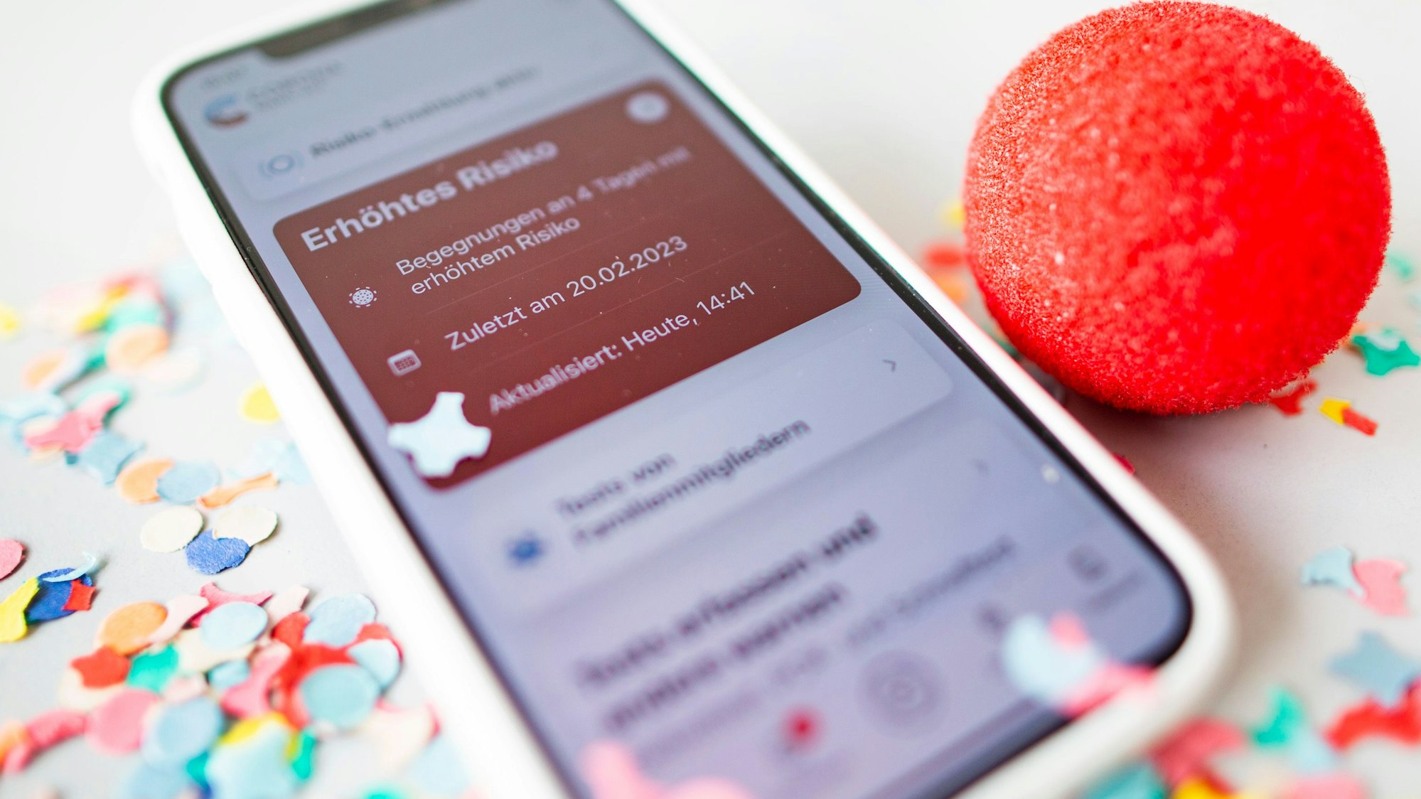 Eine Corona-Warnapp auf einem Smartphone leuchtet rot, daneben liegt Konfetti und eine Pappnase.