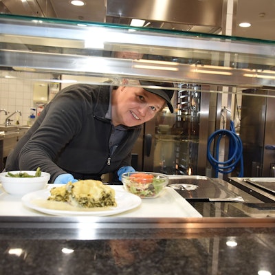 Eine Frau reicht in einer Mensa ein Essen auf einem Tablett aus der Küche heraus.