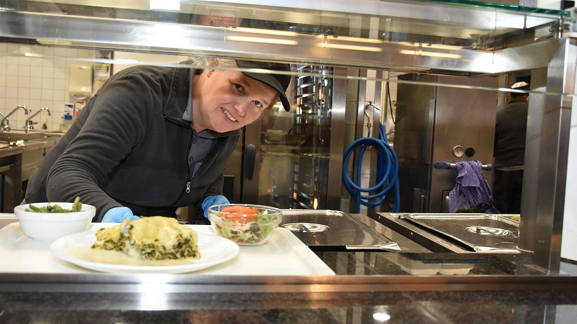 Eine Frau reicht in einer Mensa ein Essen auf einem Tablett aus der Küche heraus.
