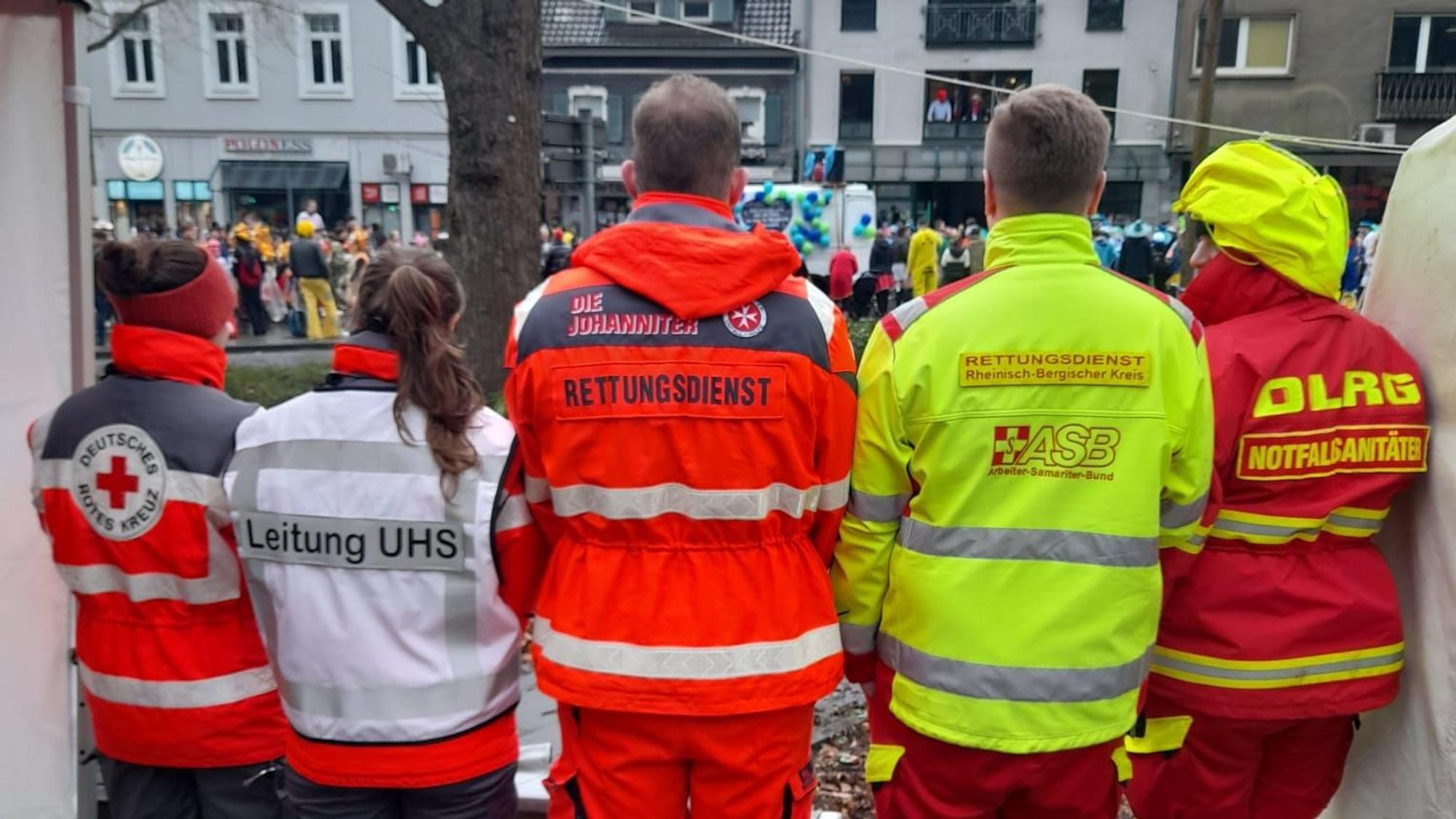 Einsatzkräfte von DRK, Johanniter, ASB und DLRG stehen nebeneinander und beobachten den Karnevalszug in Bergisch Gladbachs Stadtmitte.