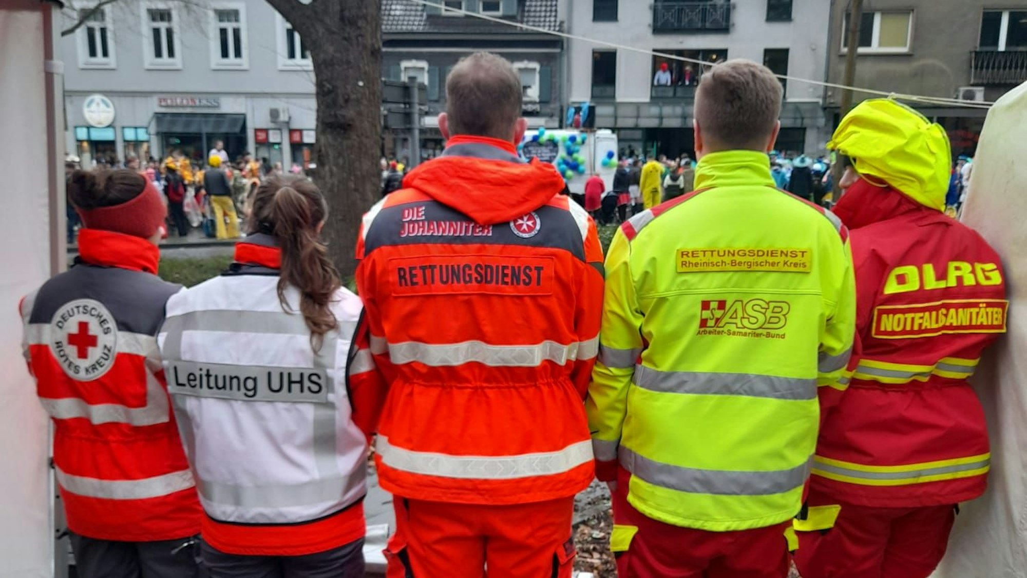 Einsatzkräfte von DRK, Johanniter, ASB und DLRG stehen nebeneinander und beobachten den Karnevalszug in Bergisch Gladbachs Stadtmitte.