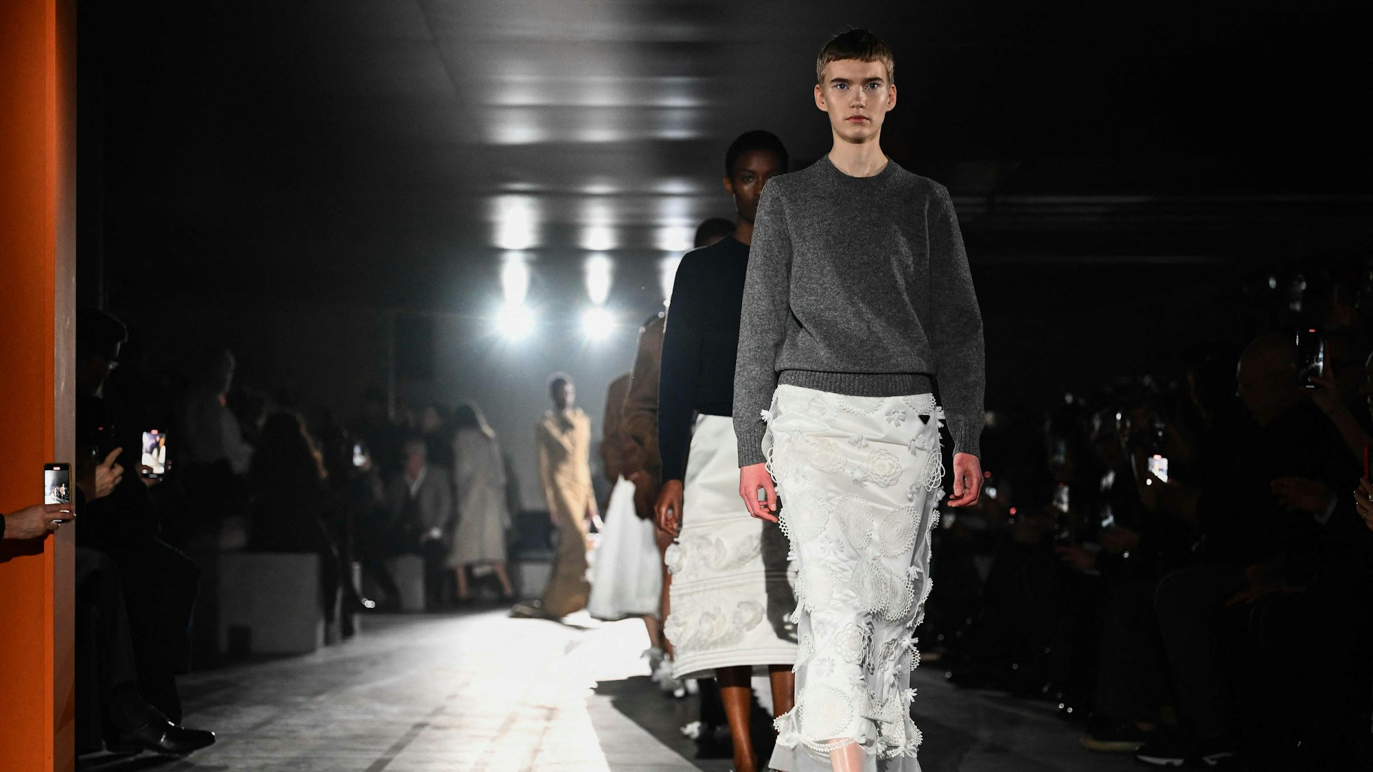 Models präsentieren Kreationen von Prada auf dem Laufsteg. Das erste Model trägt einen weißen, verschnörkelten Rock und einen schlichten grauen Pullover.