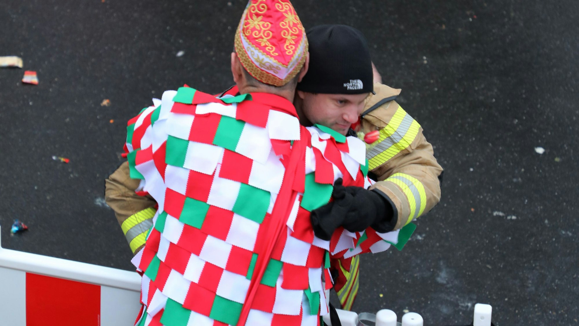 Feuerwehrchef Jörg Köhler umarmt im Lappenfrack der Großen Gladbacher KG einen Feuerwehrkollegen in Einsatzkleidung an einer Straßenabsperrung.
