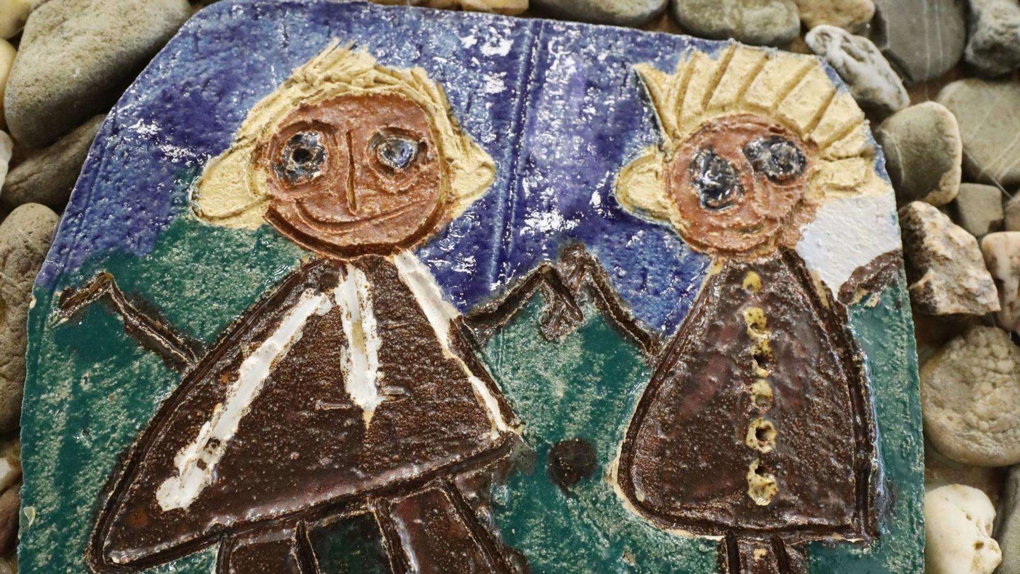 Auf einer gebrannten Keramikplatte sind vor einem grünen und blauen Hintergrund in Braun zwei Figuren dargestellt, ein Mann und eine Frau.