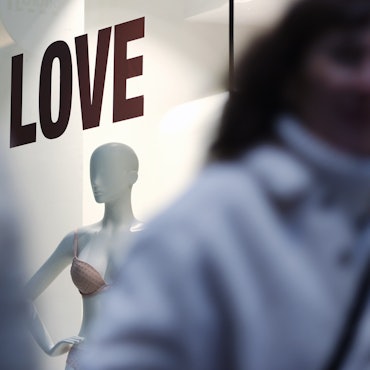 Man sieht zwischen zwei unscharfen Menschen durch fotografiert ein Schaufenster mit einer Puppe. Über ihr steht „LOVE“.