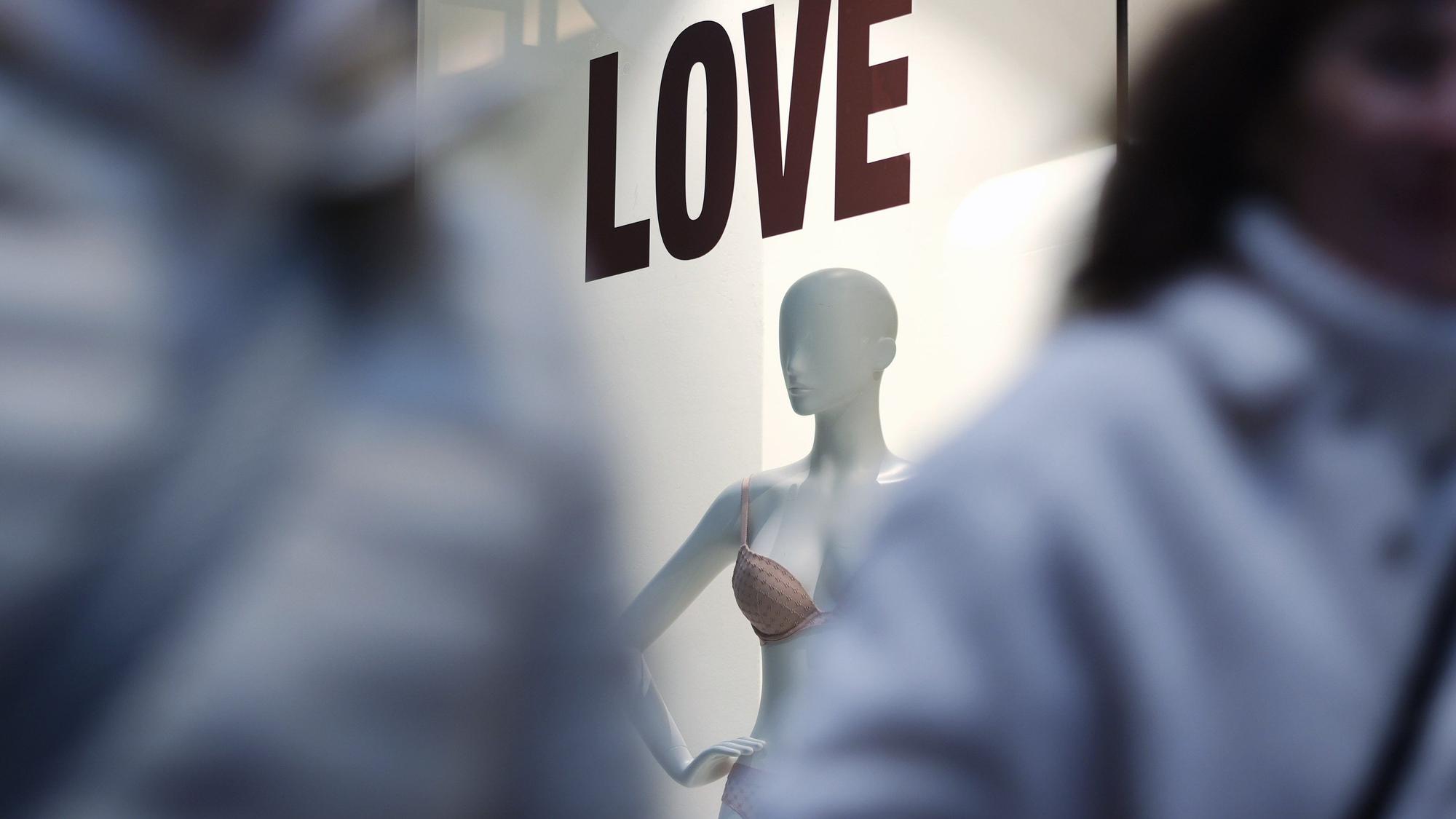 Man sieht zwischen zwei unscharfen Menschen durch fotografiert ein Schaufenster mit einer Puppe. Über ihr steht „LOVE“.