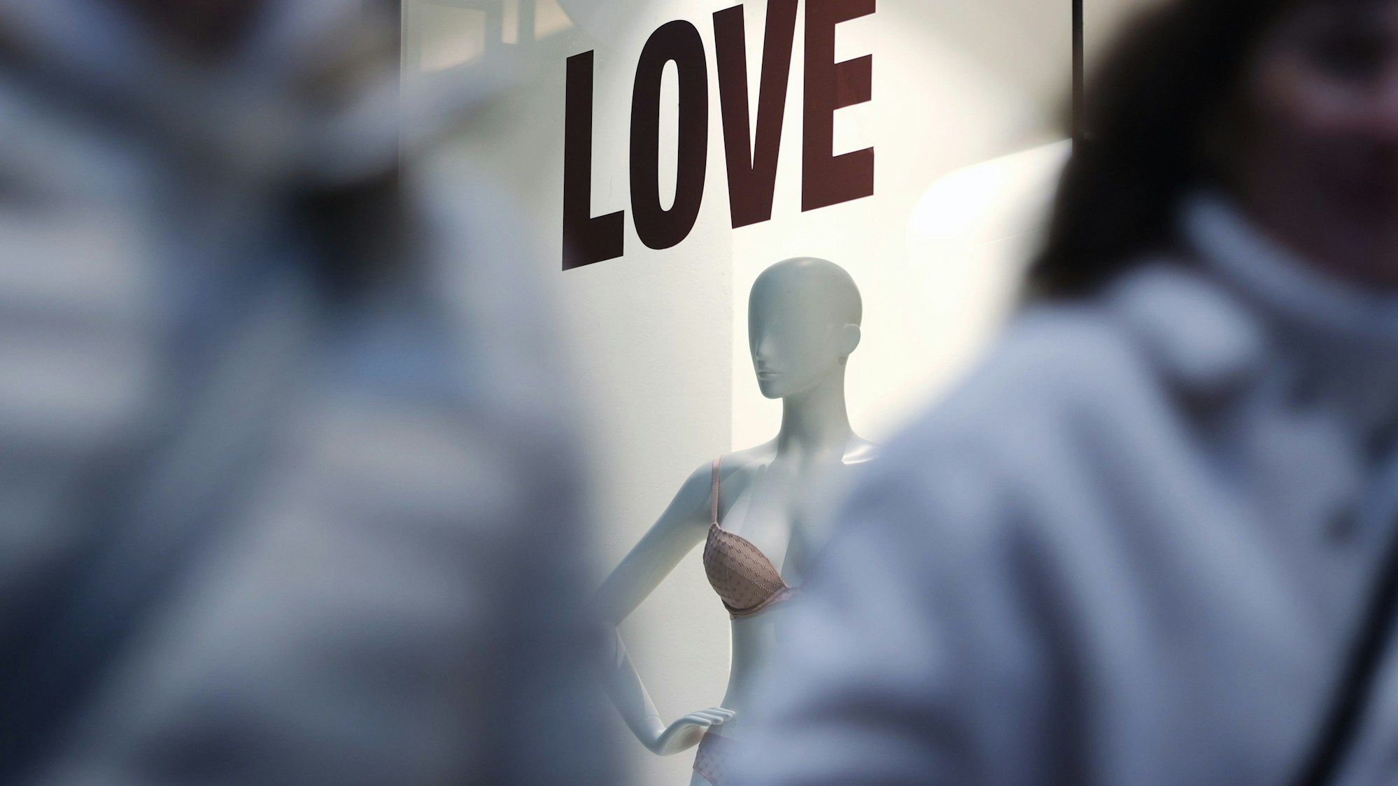Man sieht zwischen zwei unscharfen Menschen durch fotografiert ein Schaufenster mit einer Puppe. Über ihr steht „LOVE“.