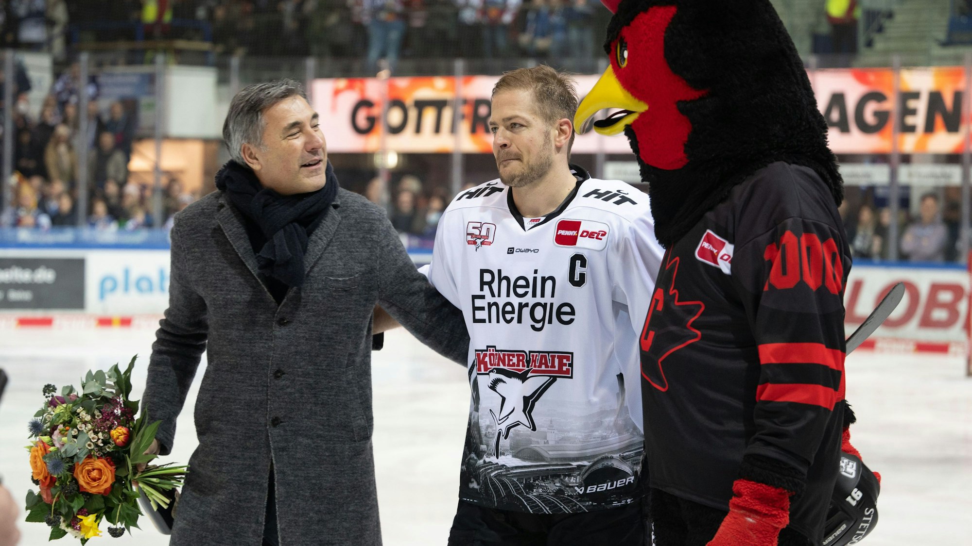 24.02.2023, Nordrhein-Westfalen, Iserlohn: Eishockey: DEL, Iserlohn Roosters - Kölner Haie, Hauptrunde, 46. Spieltag, Eissporthalle Iserlohn: Moritz Müller (M) wird vor dem Spiel für sein 1000. Spiel geehrt. Rechts Maskottchen Icey von den Rooster. Links Wolfgang Brück, Geschäftsführer der Iserlohn Roosters. Foto: Bernd Thissen/dpa +++ dpa-Bildfunk +++