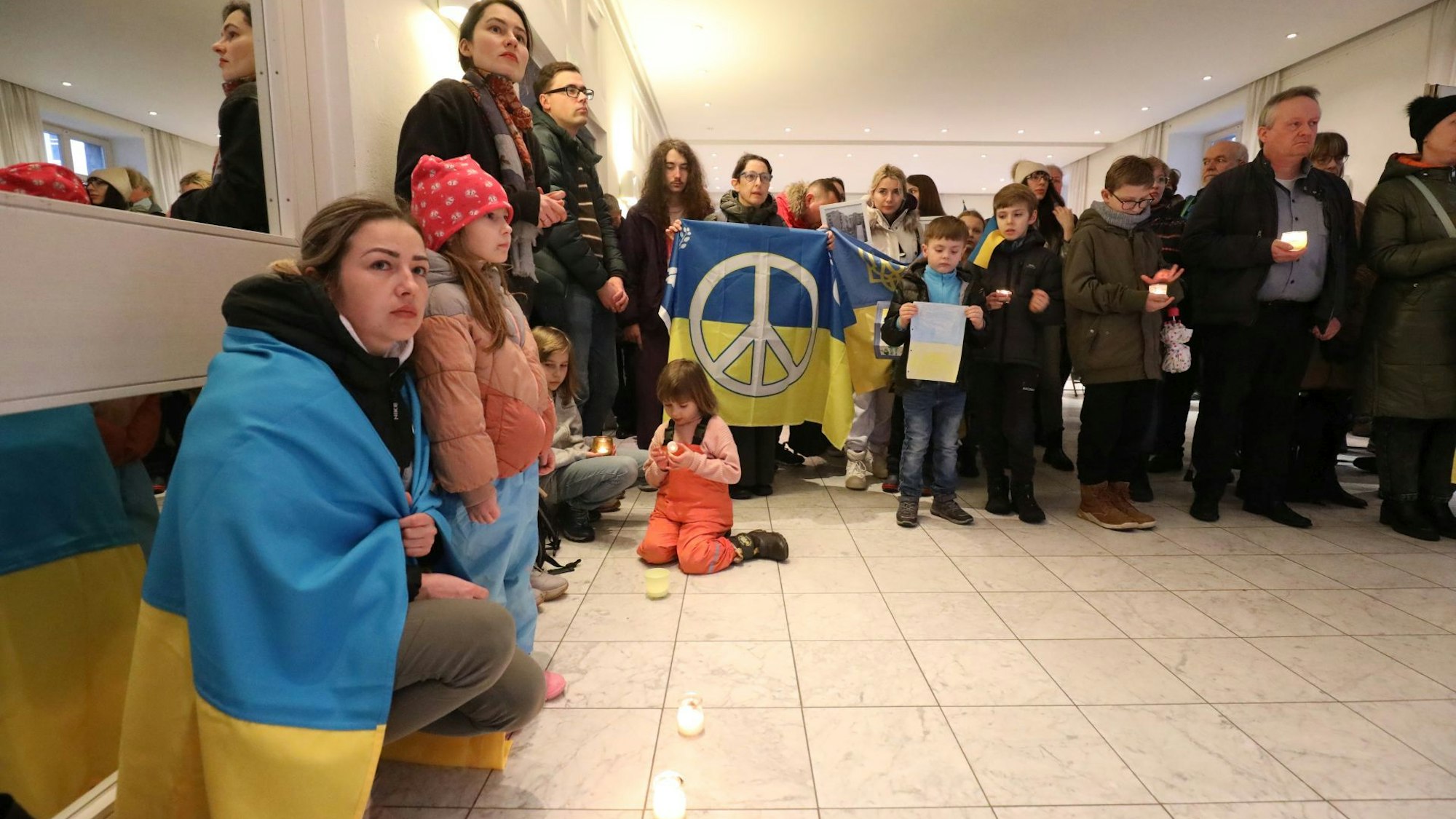 Eine Frau hat die blau-gelbe ukrainische Flagge umgehangen. Auf dem Boden stehen Windlichter. Im Hintergrund halten Menschen eine ukrainische Flagge mit dem Peace-Zeichen.