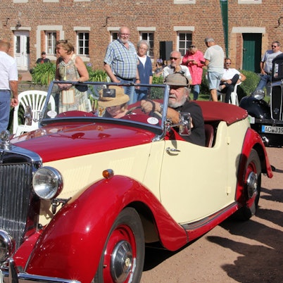 Zwei alte Autos stehen hintereinander auf einem Weg. Neben dem Weg stehen mehrere Zuschauer.