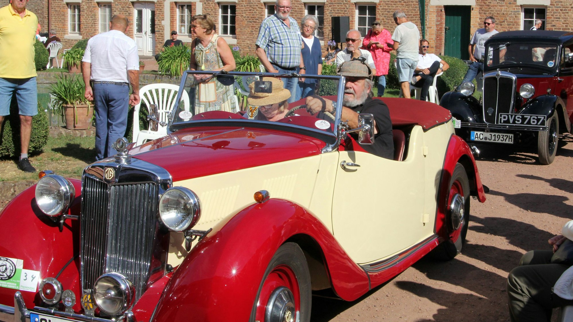 Zwei alte Autos stehen hintereinander auf einem Weg. Neben dem Weg stehen mehrere Zuschauer.