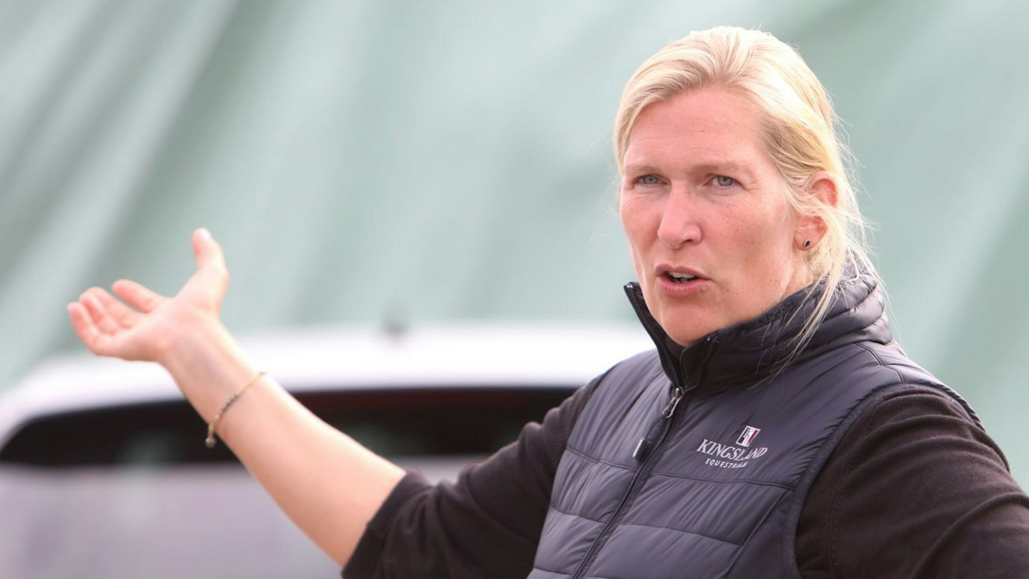 Dhana Schwarz steht mit einer blauen Weste über einem schwarzen Pullover auf ihrem Hof. Mit der recht Hand zeigt sie auf etwas.