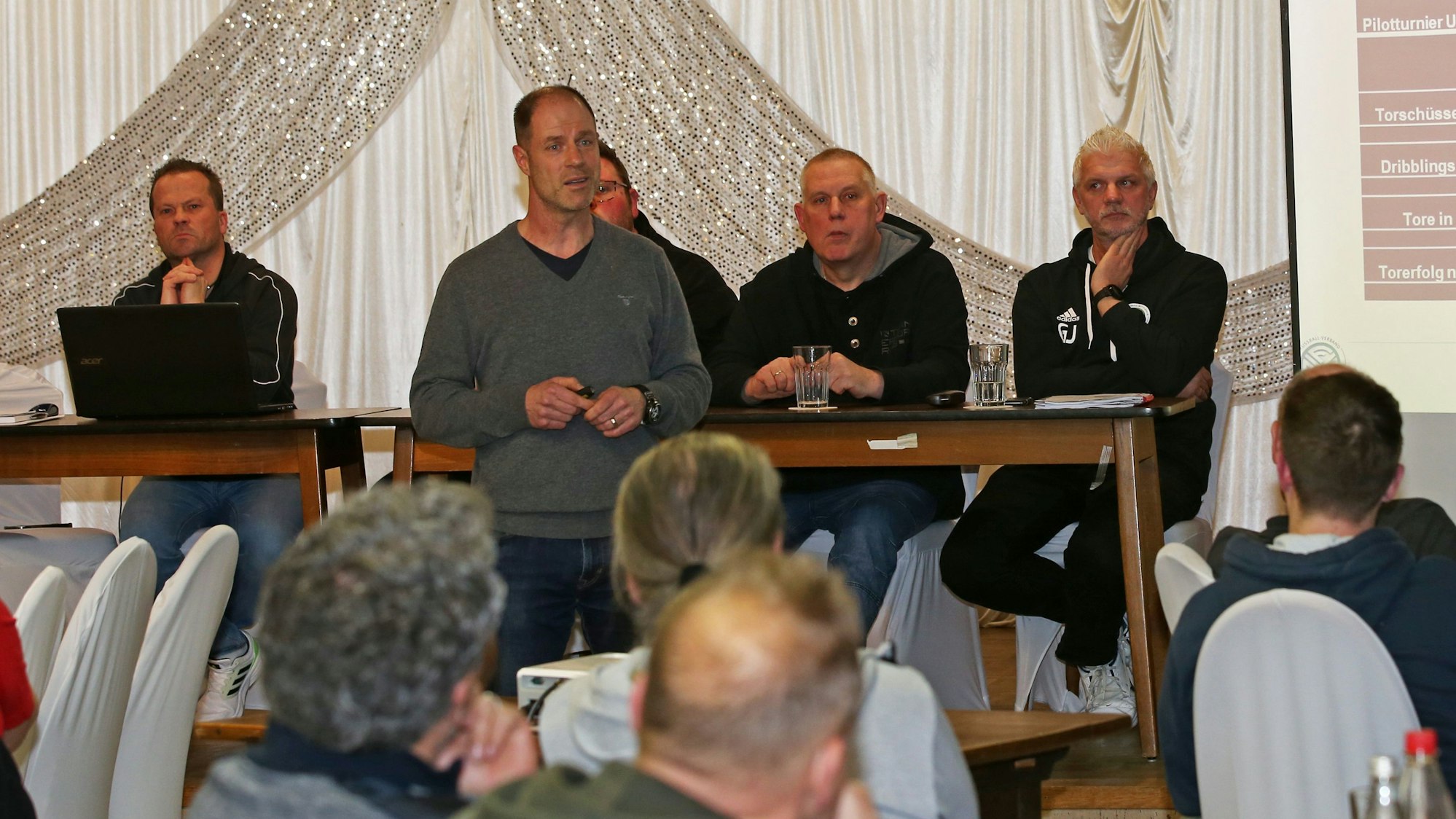 Mehrere Männer sitzen an einem Tisch. Einer steht im Vordergrund und hält einen Vortrag. Auf einer Leinwand ist eine Pärsentation zu sehen.
