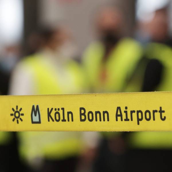 Ein gelbes Absperrband mit der Aufschrift „Köln Bonn Airport“ hängt vor eine Gruppe Streikender während eines Warnstreiks.