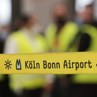 Ein gelbes Absperrband mit der Aufschrift „Köln Bonn Airport“ hängt vor eine Gruppe Streikender während eines Warnstreiks.