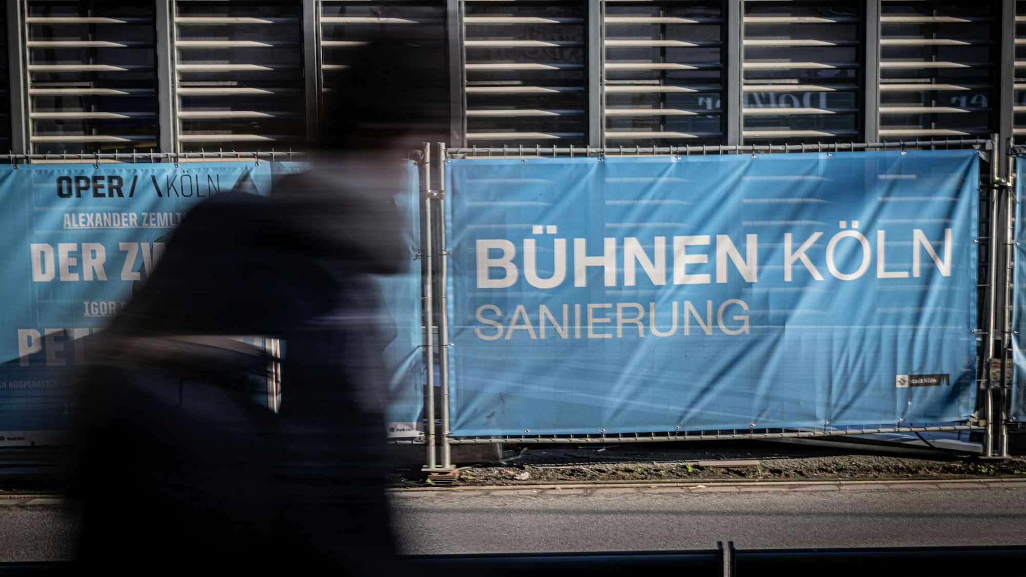 Eine verschwommene Person passiert die Baustelle. Auf dem Sperrzaun steht in weißen Buchstaben auf blauem Hintergrund: "Bühnen Köln Sanierung".