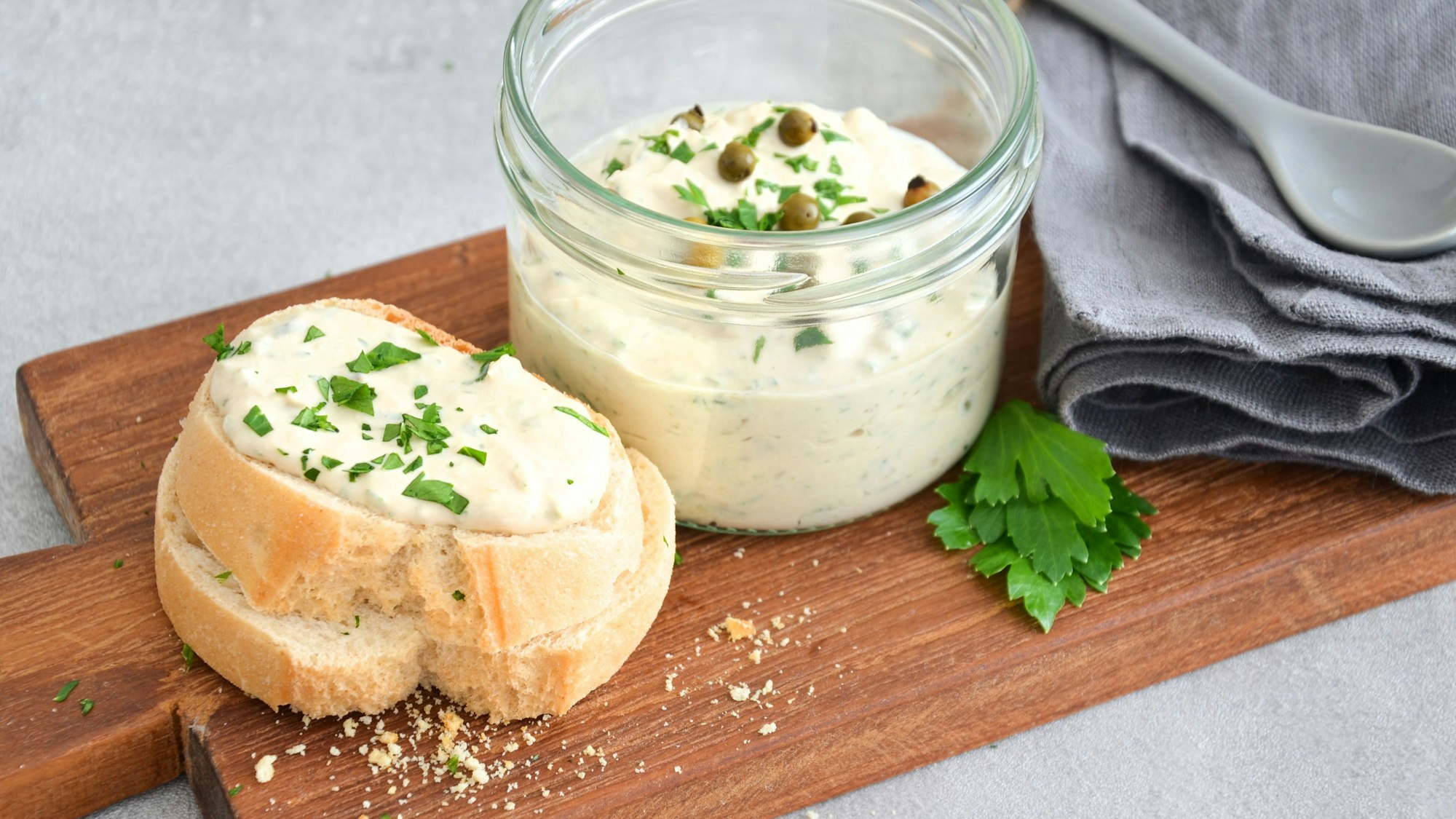 Ein Thunfisch-Mascarponedip angerichtet auf einem Holzbrettchen mit Baguette Brot und grünem Pfeffer.