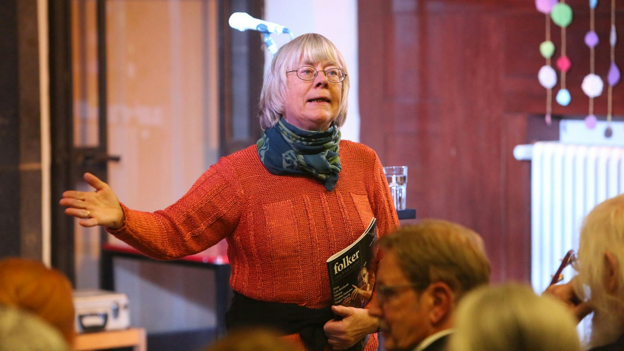 Eine Frau im roten Pullover spricht vor einer Gruppe von Konzertbesuchern, in der Hand hält sie das Magazin „Folker“