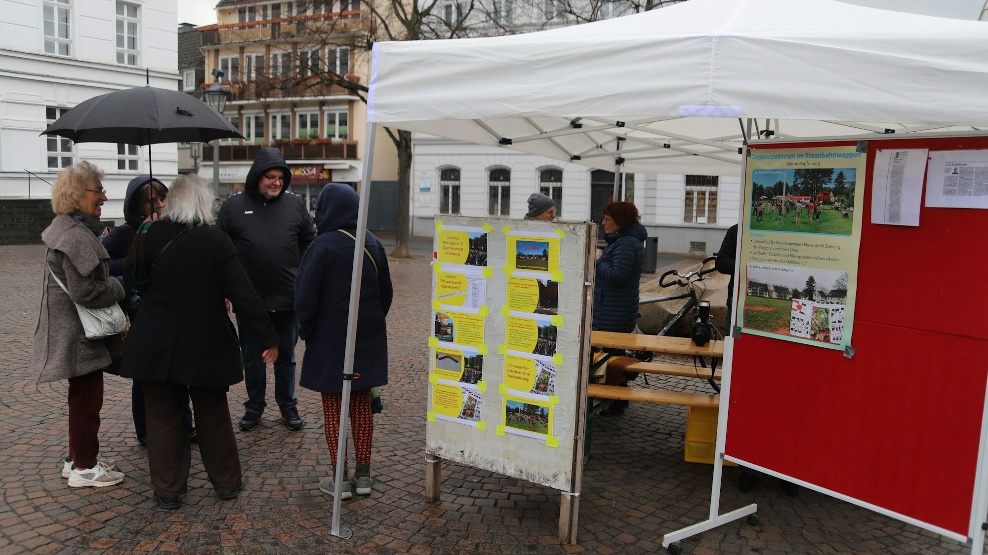 Blick auf das weiße Infostand-Zelt. Einige Leute stehen auf dem Platz davor.