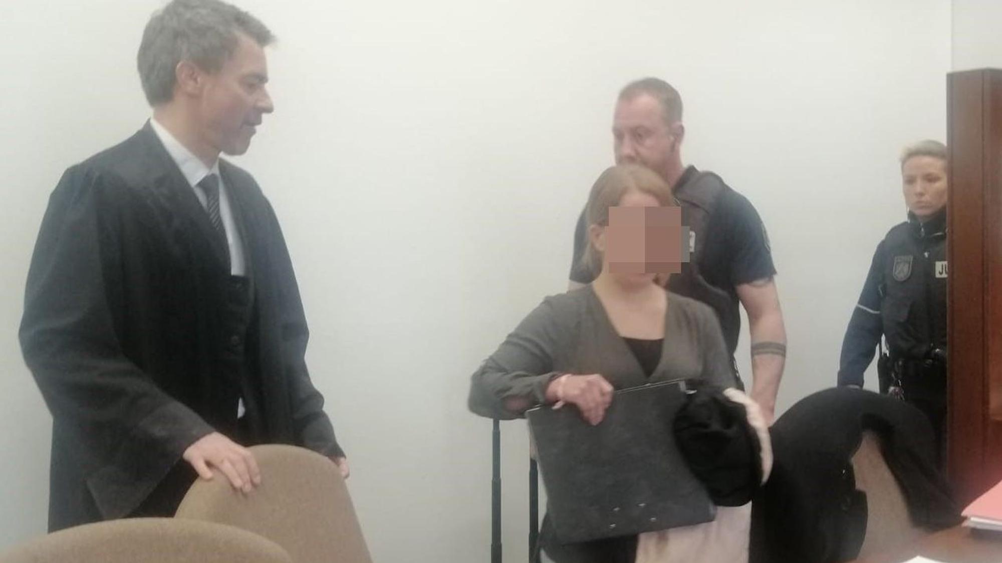 Das Foto zeigt die Angeklagte im Gerichtssaal. Zu sehen sind zudem ihr Verteidiger und zwei Justizvollzugsbeamte.