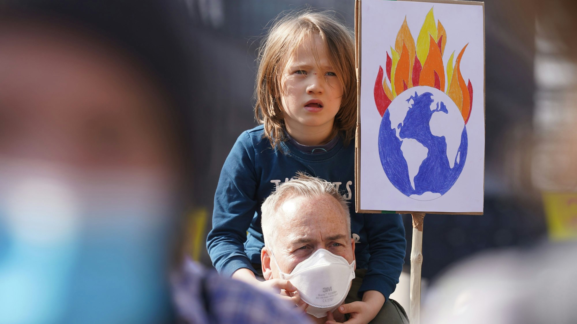 Emil sitzt auf den Schultern vom Vater Oli zwischen den Teilnehmern des Klimastreiks Fridays for Future. Der Vater hält ein Plakat mit einer brennenden Erde hoch.