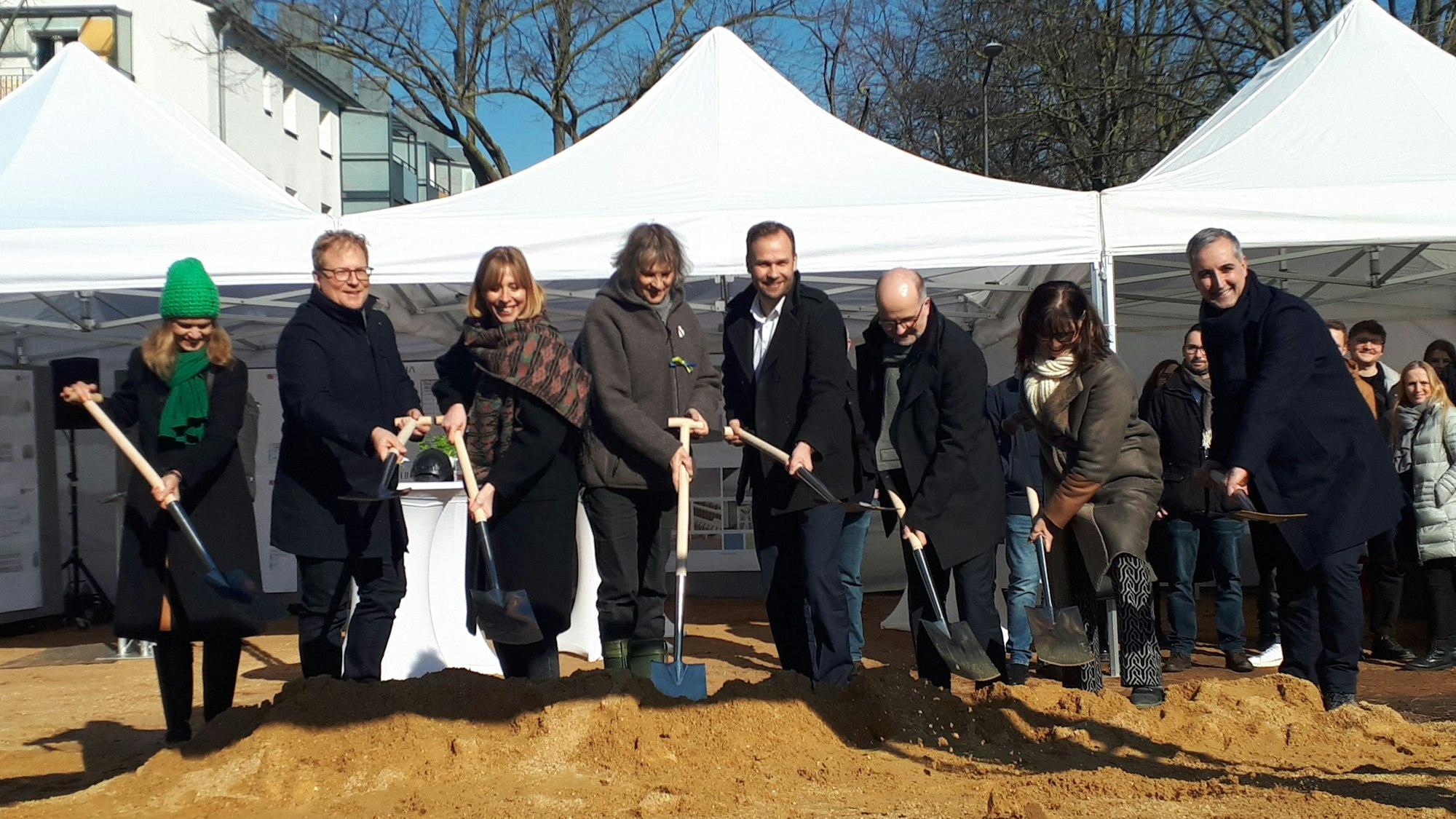 Acht Menschen stehen mit Spaten nebeneinander, sie stehen auf einem Baudgrundstück mit sandigem Boden, hinter ihnen sind weiße Pavillons zu sehen.