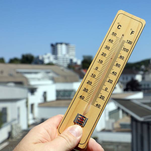 Jemand hält ein Thermometer in Bergisch Gladbach vor den strahlend blauen Himmel und die Häuserkulisse im Hintergrund.