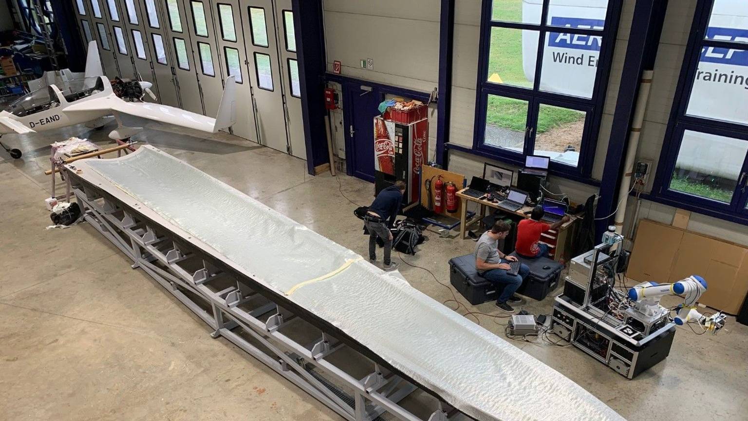 Bei der Aeroconcept GmbH ist für einen Test ein Faserpaket für eine Rotorblattspitze ausgelegt worden. Techniker sitzen neben der Form in einer Flugzeughalle.