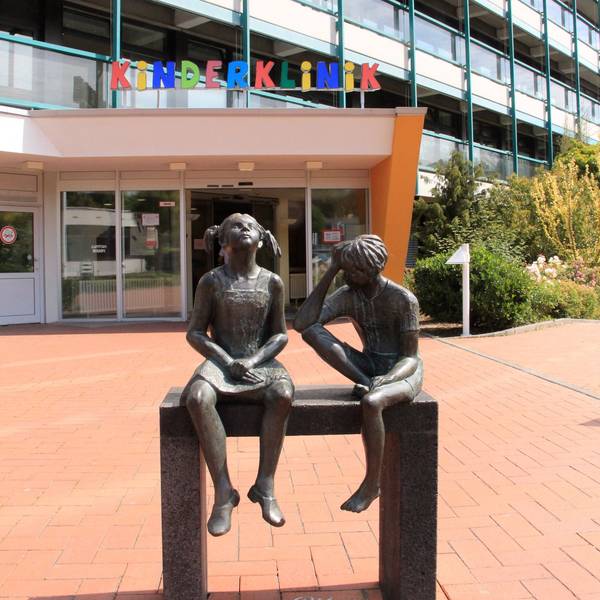 Über dem Eingang steht in bunten Buchstaben Kinderklinik. Im Vordergrund sind zwei Statuen von Kindern zu sehen.