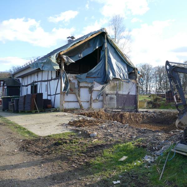 Ein provisorisch abgedecktes Wohnhaus auf Hof Huppenhardt, rechts davor steht ein Bagger und eine Palette mit Dachziegeln.
