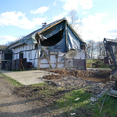 Ein provisorisch abgedecktes Wohnhaus auf Hof Huppenhardt, rechts davor steht ein Bagger und eine Palette mit Dachziegeln.
