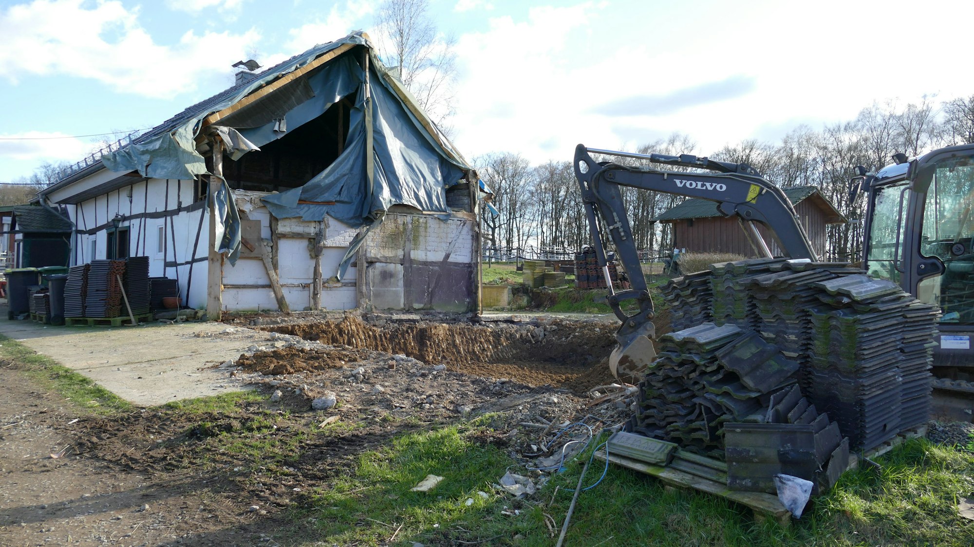 Ein provisorisch abgedecktes Wohnhaus auf Hof Huppenhardt, rechts davor steht ein Bagger und eine Palette mit Dachziegeln.
