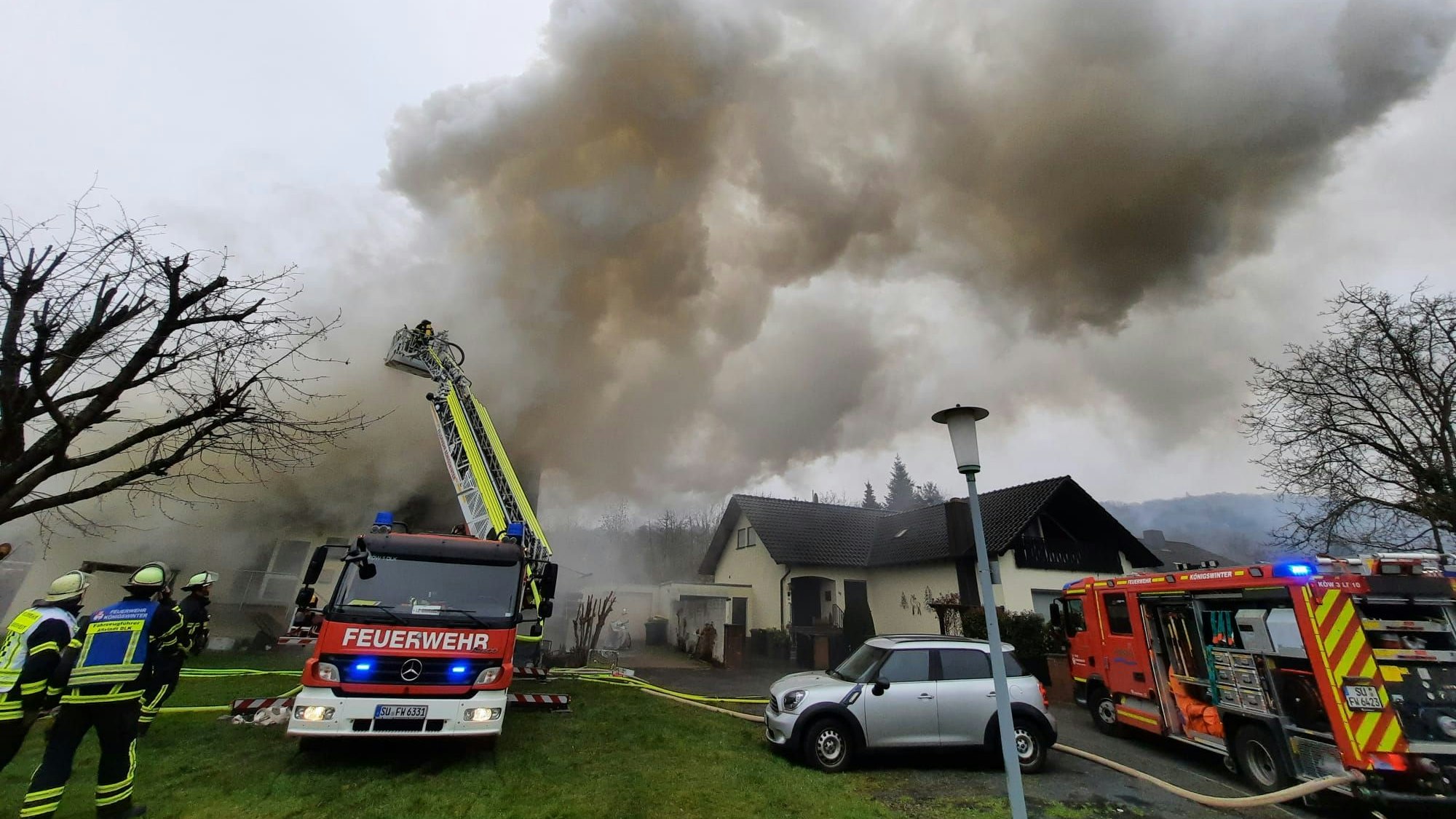 Über einem Haus steigt eine dichte, graue Rauchwolke auf. Von einer Drehleiter aus löschen Feuerwehrleute den brennenden Dachstuhl..