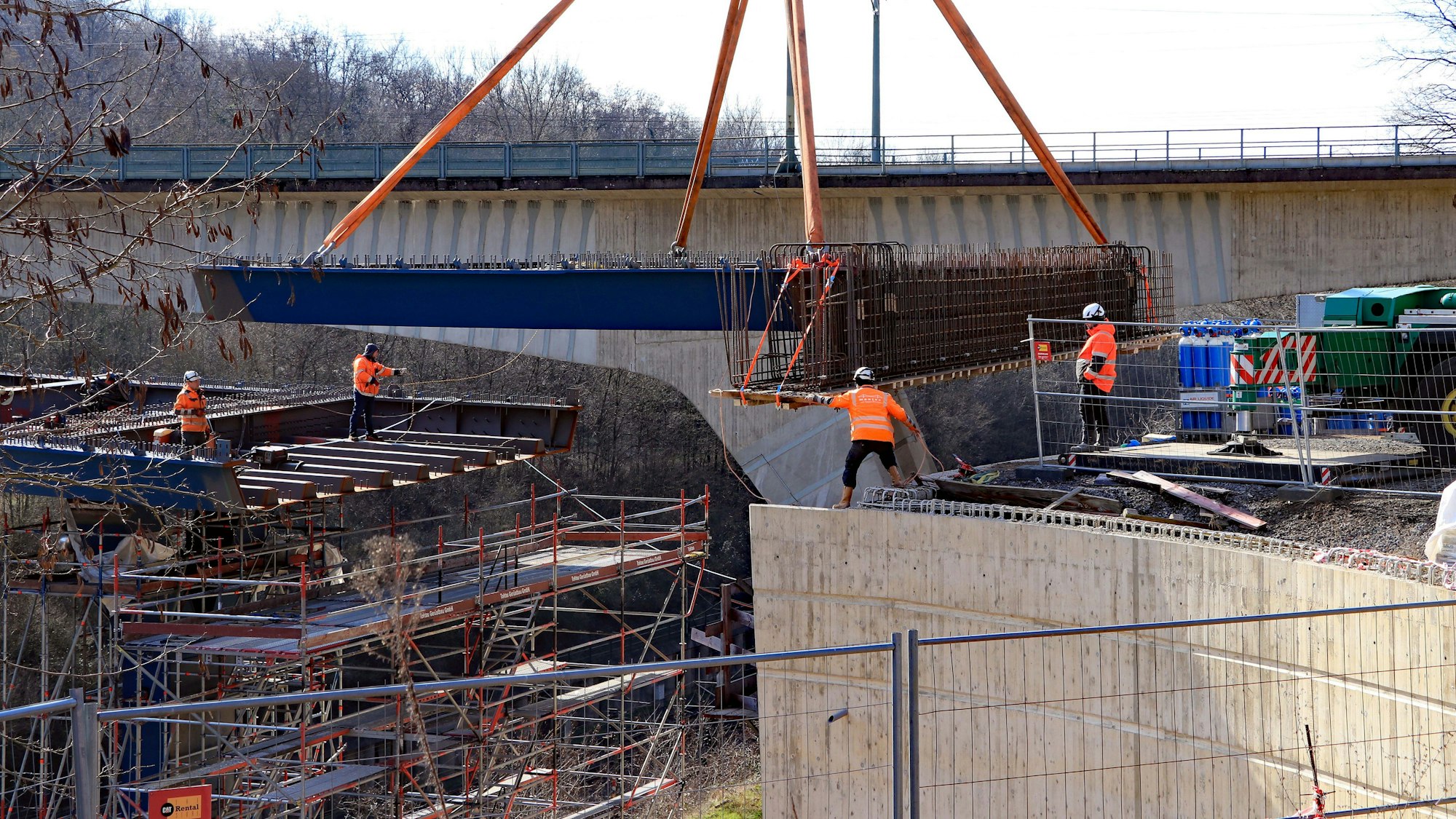 Auf dem Foto ist zu sehen, wie der Schwerlastkran das tonnenschwere Brückenelement in seine endgültige Position hievt. Mehrere Bauarbeiter helfen dabei.
