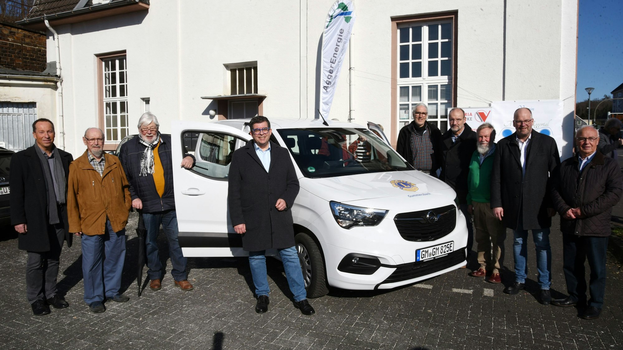 Neun Männer stehen rund um ein neues, weißes Elektroauto.