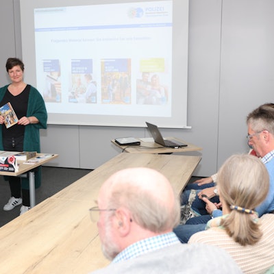 Eine Frau hält einen Vortrag. Einige ältere Besucher hören ihr zu.